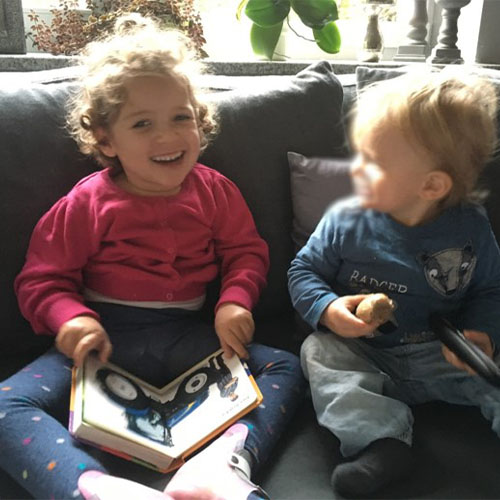 Zwei kleine Kinder sitzen auf der Couch. Das Mädchen lacht in die Kamera und hat ein aufgeschlagenes Buch im Schoß, in dem ein Traktor abgebildet ist. Der kleinere Junge daneben schaut sie an.