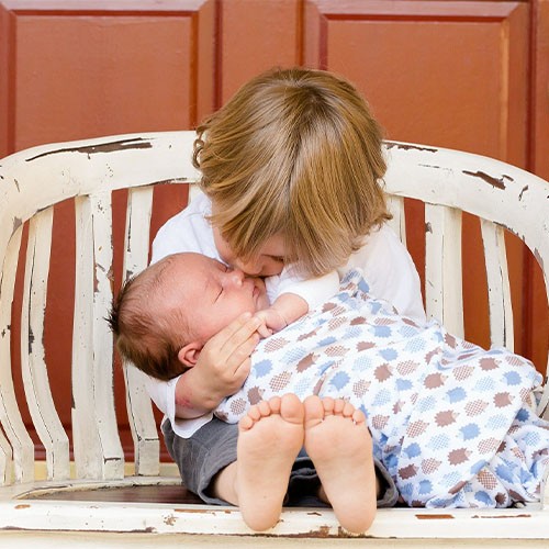 Kleinkind sitzt auf einer weißen Bank und hält ein schlafendes Baby im Arm, das es zärtlich auf die Wange küsst.