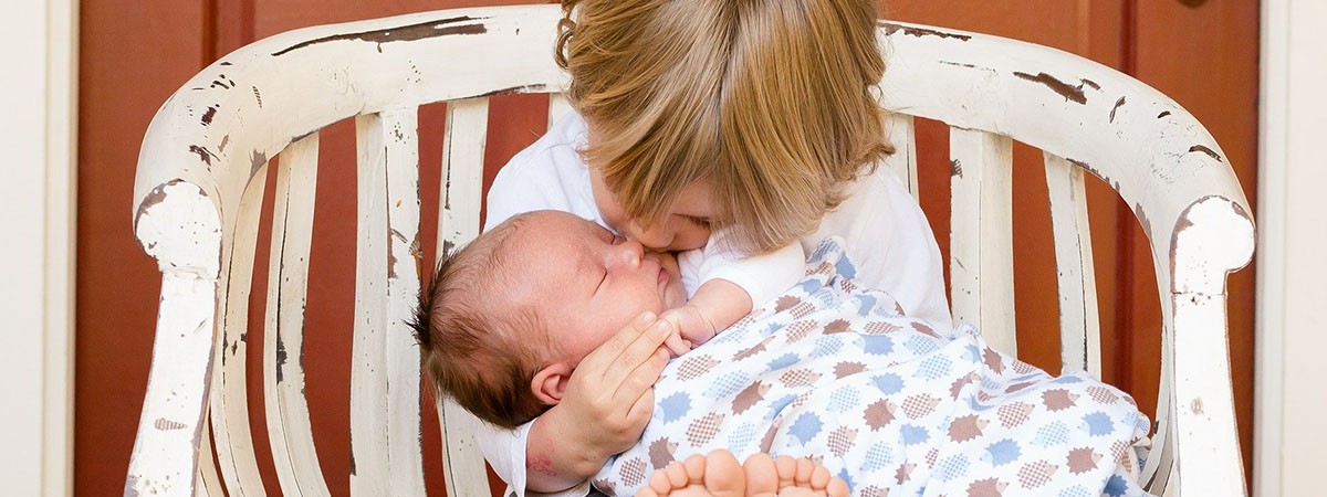 Kleinkind sitzt auf einer weißen Bank und hält ein schlafendes Baby im Arm, das es zärtlich auf die Wange küsst.
