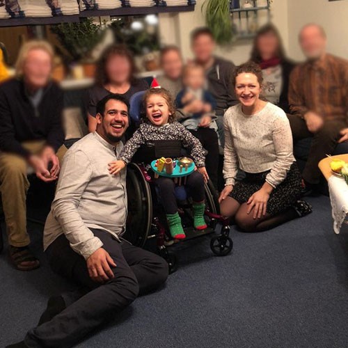 Familienportrait: Auf dem Bild sind mehrere Familienmitglieder zu sehen. Darunter auch die kleine SMA-Patientin und ihre Eltern. Die Personen sitzen im Wohnzimmer auf dem Sofa und dem Boden und lachen in die Kamera.