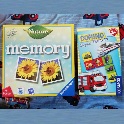 Zwei Kartons von Kinderspielen liegen auf einer Kinderdecke. Das eine ist ein Memory-, das andere ein Domino-Spiel.