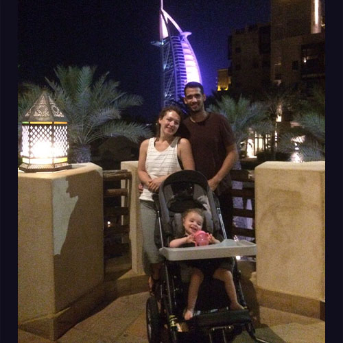 Das Urlaubsbild zeigt die kleine SMA-Patientin in ihrem Rehabuggy und die beiden Eltern, die Arm in Arm hinter dem Mädchen stehen. Im Hintergrund ist das nächtliche Dubai.