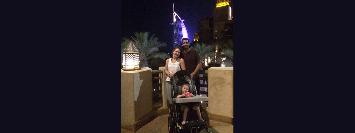 Das Urlaubsbild zeigt die kleine SMA-Patientin in ihrem Rehabuggy und die beiden Eltern, die Arm in Arm hinter dem Mädchen stehen. Im Hintergrund ist das nächtliche Dubai.