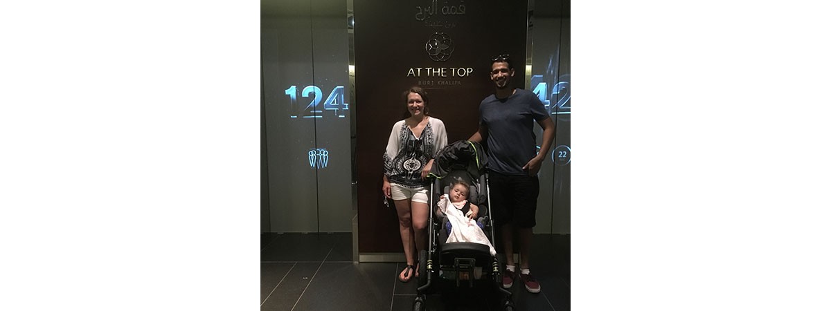 Vater und Mutter stehen im Urlaub in Dubai in einem Gebäude vor den Aufzügen. Vor ihnen steht der Regabuggy, in dem ihre Tochter sitzt und hinter ihnen steht „AT THE TOP, BURJ KHALIFA“ an der Wand.