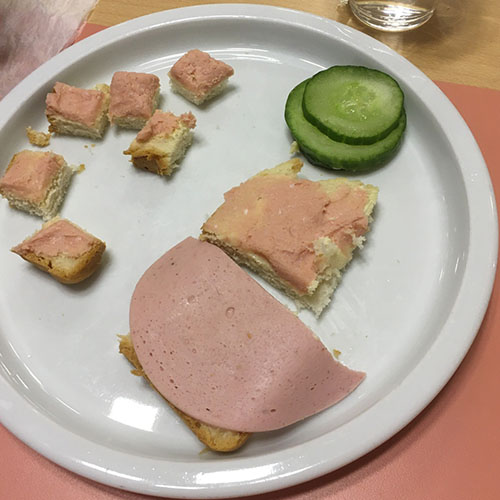 Auf einem Teller liegen Gurkenscheiben sowie große und kleine Stücke Brot, die belegt sind mit Fleischwurst und einer Streichwurst.