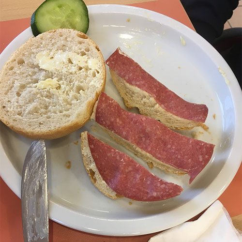 Auf einem Teller liegen eine Brötchenhälfte sowie eine in drei Streifen geschnittene Brötchenhälfte, die mit einer Salamischeibe belegt ist.