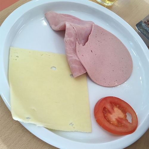 Auf einem Teller liegen eine Scheibe Käse, eine angebissene Scheibe Fleischwurst sowie Kochschinken und eine Tomatenscheibe.