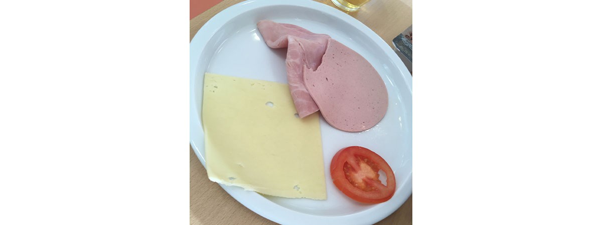 Auf einem Teller liegen eine Scheibe Käse, eine angebissene Scheibe Fleischwurst sowie Kochschinken und eine Tomatenscheibe.