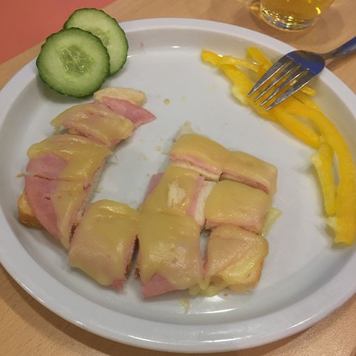 Auf einem Teller liegen ein in kleine Stückchen geschnittenes Toastbrot mit Wurst belegt und Käse überbacken sowie zwei Scheiben Gurke und Streifen von gelber Paprika.