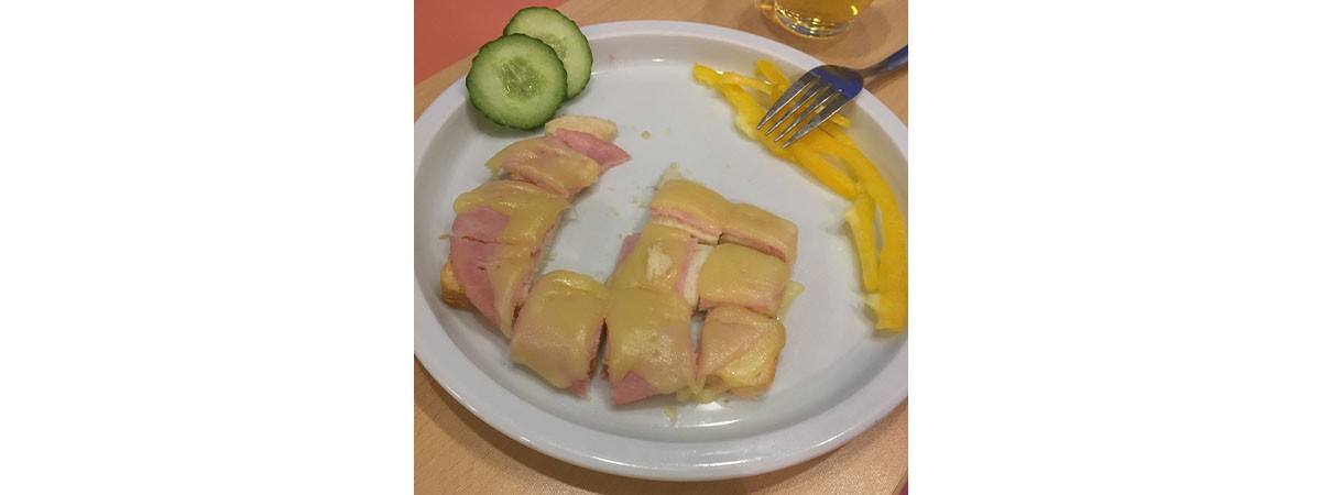 Auf einem Teller liegen ein in kleine Stückchen geschnittenes Toastbrot mit Wurst belegt und Käse überbacken sowie zwei Scheiben Gurke und Streifen von gelber Paprika.