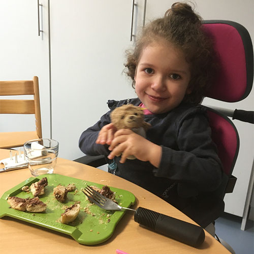 Kleine SMA-Patientin ist in der Reha und sitzt am Tisch. Vor ihr ist ein grünes Brett mit Brotstückchen und einer therapeutischen Gabel. Sie lächelt und hält einen Teddy in den Händen.