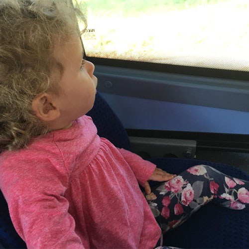 Blondes Kleinkind (SMA-Patientin) mit lockigem Haar und einem pinken Shirt schaut aus dem Fenster eines Zuges.
