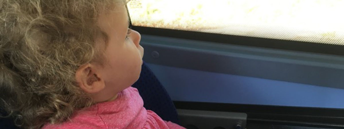 Blondes Kleinkind (SMA-Patientin) mit lockigem Haar und einem pinken Shirt schaut aus dem Fenster eines Zuges.