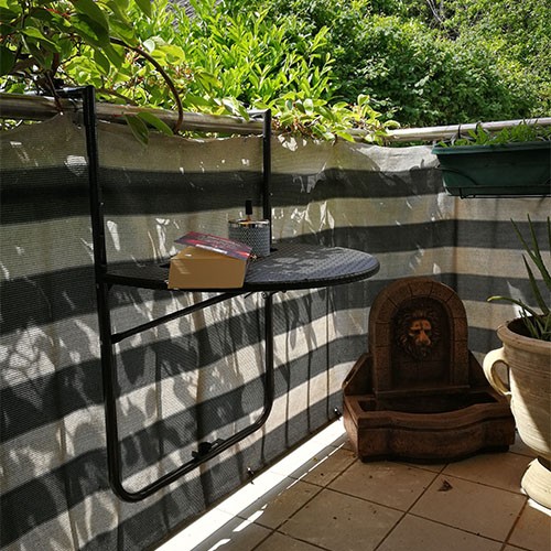 Ein Balkon mit gestreiftem Sichtschutz. An dem Geländer hängt ein halbrunder Tisch, auf dem ein Buch liegt. In der Ecke steht ein kleiner Brunnen mit Löwenkopf. Im Hintergrund sind Büsche und Baumkronen.