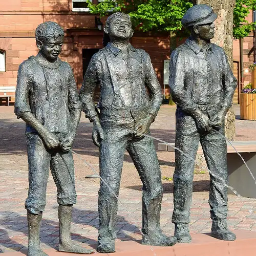 Drei männliche Figuren lassen Wasser. Die Statuen stehen auf einem Platz in der Öffentlichkeit und pinkeln Wasser.