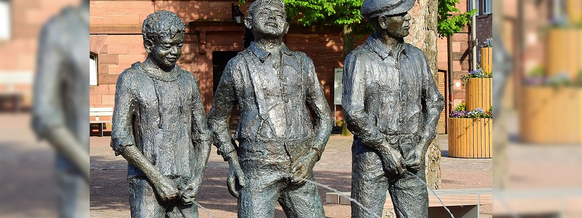 Drei männliche Figuren lassen Wasser. Die Statuen stehen auf einem Platz in der Öffentlichkeit und pinkeln Wasser.