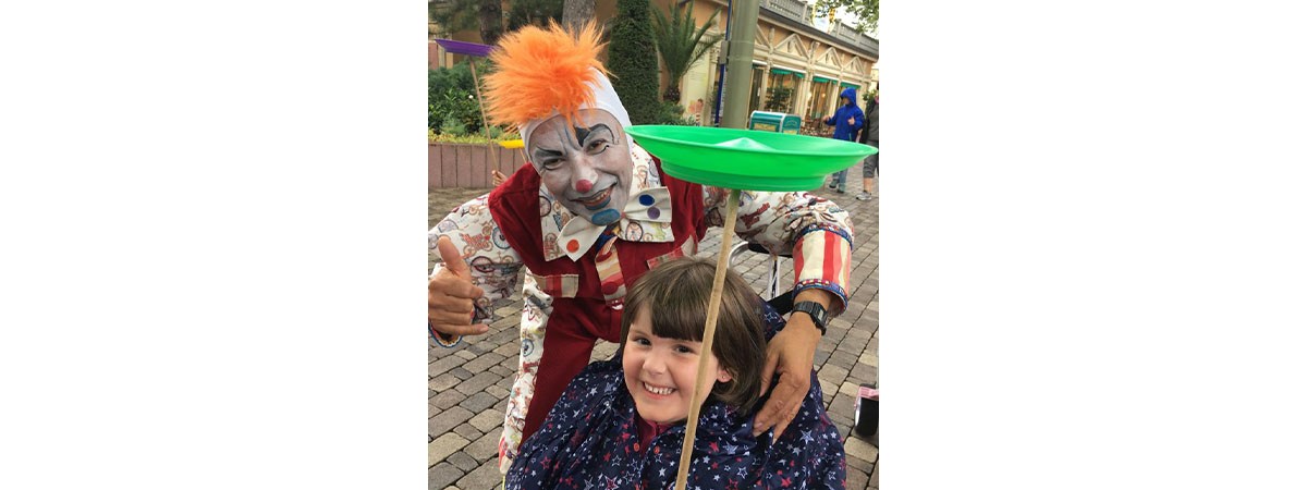 Die kleine SMA-Patientin lächelt in die Kamera. Hinter ihr steht ein Clown des Freizeitparks.