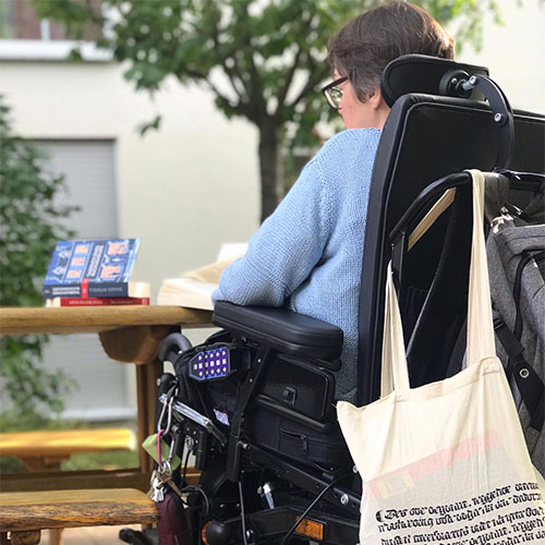 Patientin sitzt mit dem Rücken zum Betrachter in einem Rollstuhl am Tisch und liest ein Buch.