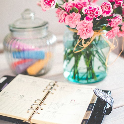 Ein Terminkalender zum Planen des Alltags sowie privater oder beruflicher Termine, ein Glasbehälter mit Kreide und ein Strauß Blumen stehen auf einem Tisch.