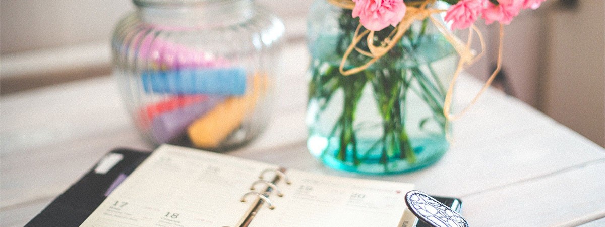 Ein Terminkalender zum Planen des Alltags sowie privater oder beruflicher Termine, ein Glasbehälter mit Kreide und ein Strauß Blumen stehen auf einem Tisch.