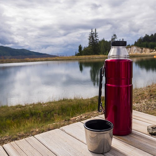 Eine rote Thermoskanne und der passende Deckel aus Edelstahl stehen auf einem Holztisch in der freien Natur. Im Hintergrund ist ein See zu sehen.