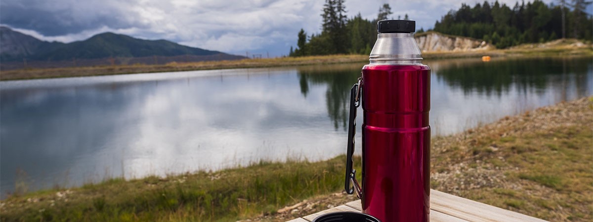 Eine rote Thermoskanne und der passende Deckel aus Edelstahl stehen auf einem Holztisch in der freien Natur. Im Hintergrund ist ein See zu sehen.