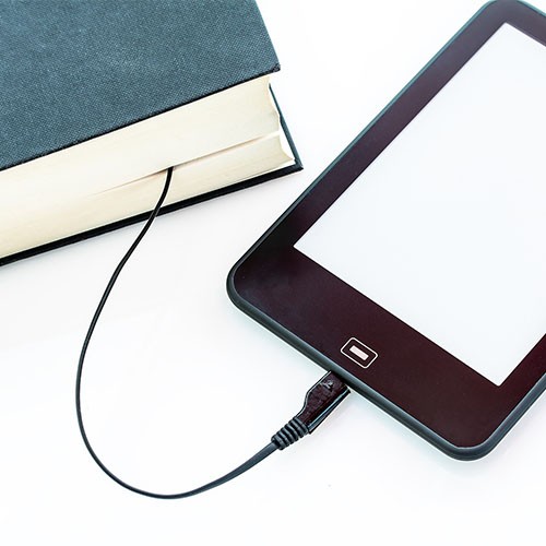 Ein Tablet oder E-Reader wird mit einem Ladekabel geladen. Das Ende des Kabels steckt in einem Buch.