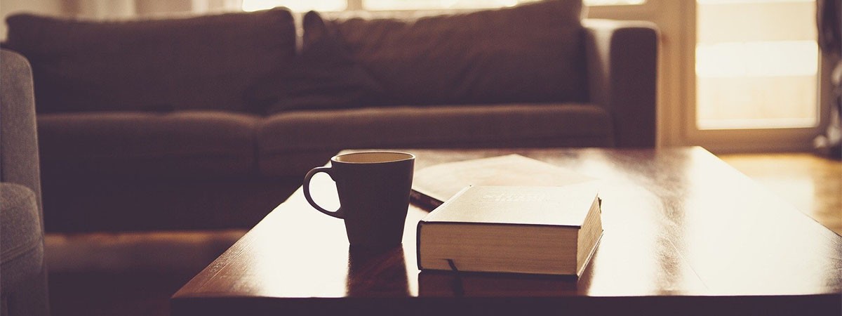 Auf einem Couchtisch im Wohnzimmer befinden sich Bücher und eine Kaffeetasse. Dahinter ist ein Sofa zu sehen. Die Sonne scheint durch die hellen Fenster und die Balkontür.