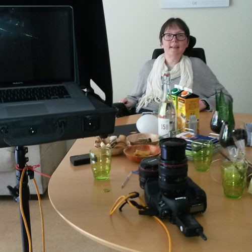 SMA-Patientin sitzt in ihrem Rollstuhl am Tisch in ihrer Wohnung. Auf ihrem Esstisch liegen eine Kamera sowie anderes Equipment, daneben steht ein Stativ mit Laptop.