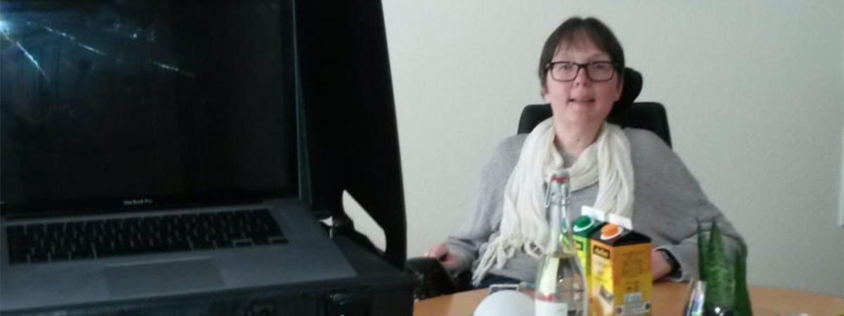 SMA-Patientin sitzt in ihrem Rollstuhl am Tisch in ihrer Wohnung. Auf ihrem Esstisch liegen eine Kamera sowie anderes Equipment, daneben steht ein Stativ mit Laptop.