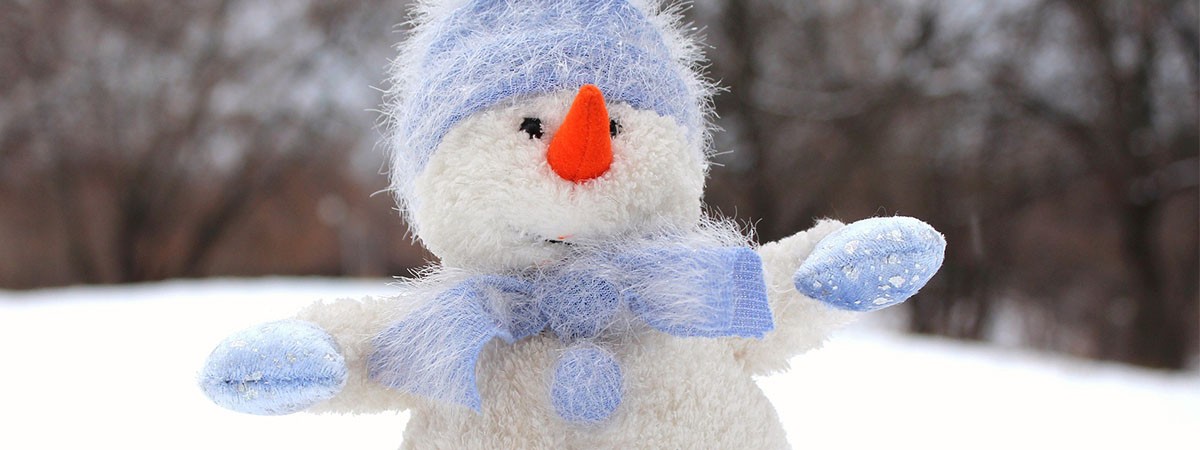 Ein Schneemann aus Stoff steht im Winter im Schnee und hat eine blaue Pudelmütze, Handschuhe und einen Schal an.