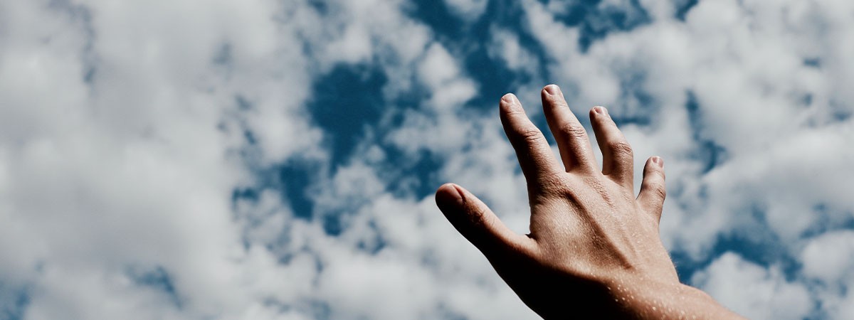 Eine Hand greift in die Luft als wolle sie nach Träumen greifen wollen. Im blauen Himmel ziehen weiße Wolken.