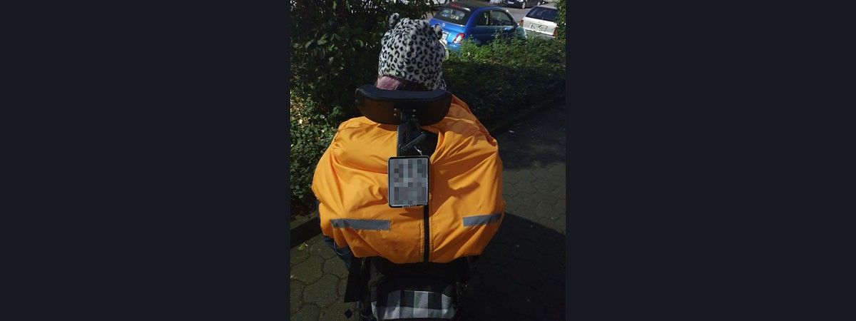 Eine SMA-Patientin sitzt draußen im Rollstuhl mit dem Rücken zur Kamera. Sie trägt Winterkleidung: eine warme Mütze sowie eine spezielle Daunenjacke für Rollstuhlfahrer.