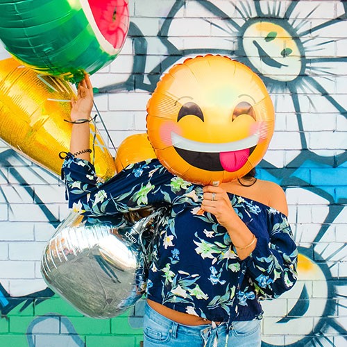Eine Frau steht vor einer mit Blumen verzierten Wand und trägt mehrere bunte Luftballons bei sich. Einen gelben Smiley-Luftballon hält sie sich vor ihr Gesicht. Der Emoji lacht fröhlich und streckt dabei frech die Zunge heraus.