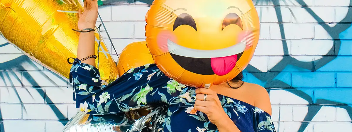 Eine Frau steht vor einer mit Blumen verzierten Wand und trägt mehrere bunte Luftballons bei sich. Einen gelben Smiley-Luftballon hält sie sich vor ihr Gesicht. Der Emoji lacht fröhlich und streckt dabei frech die Zunge heraus.