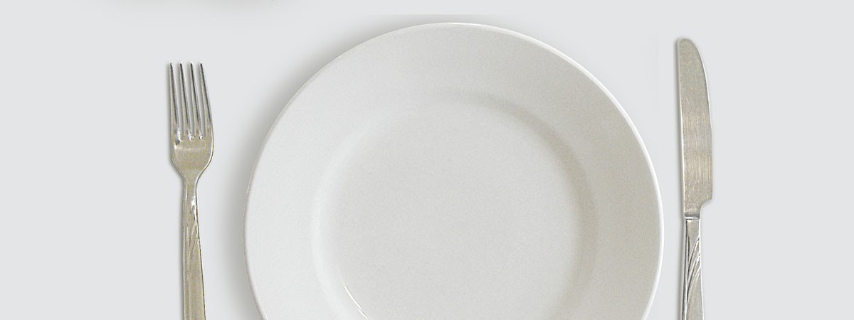 Ein leerer Teller, Besteck und ein Glas stehen auf einem weißen Esstisch.