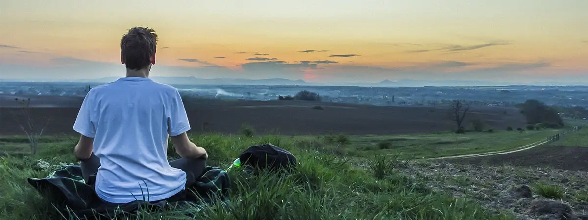Ein Mann mit weißem T-Shirt sitzt auf einer grünen Wiese und schaut in Richtung Horizont, wo der Himmel sich im Sonnenauf- oder Untergang verfärbt.