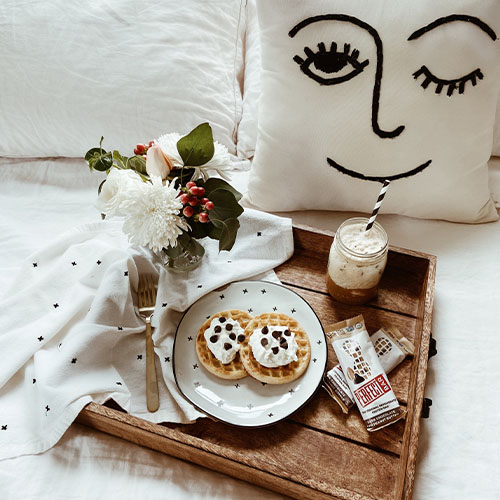 Frühstück im Bett: Auf einem Holztablett stehen Riegel, Pfannkuchen, ein Getränk und ein Blumenstrauß. Dahinter ist ein weißes Kissen mit einem lächelnden, zwinkernden Gesicht.