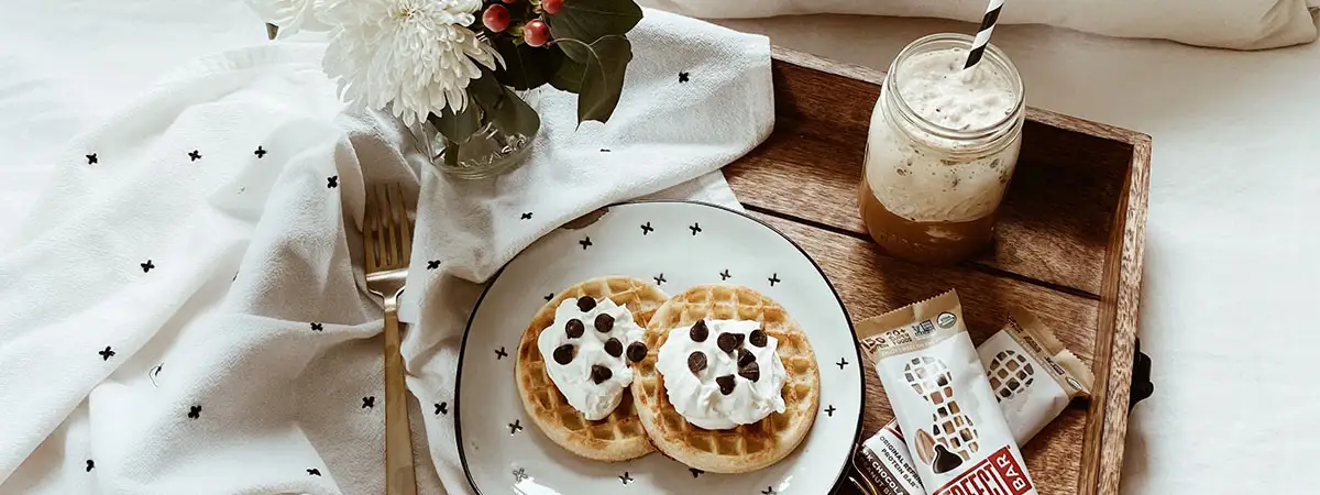 Frühstück im Bett: Auf einem Holztablett stehen Riegel, Pfannkuchen, ein Getränk und ein Blumenstrauß. Dahinter ist ein weißes Kissen mit einem lächelnden, zwinkernden Gesicht.