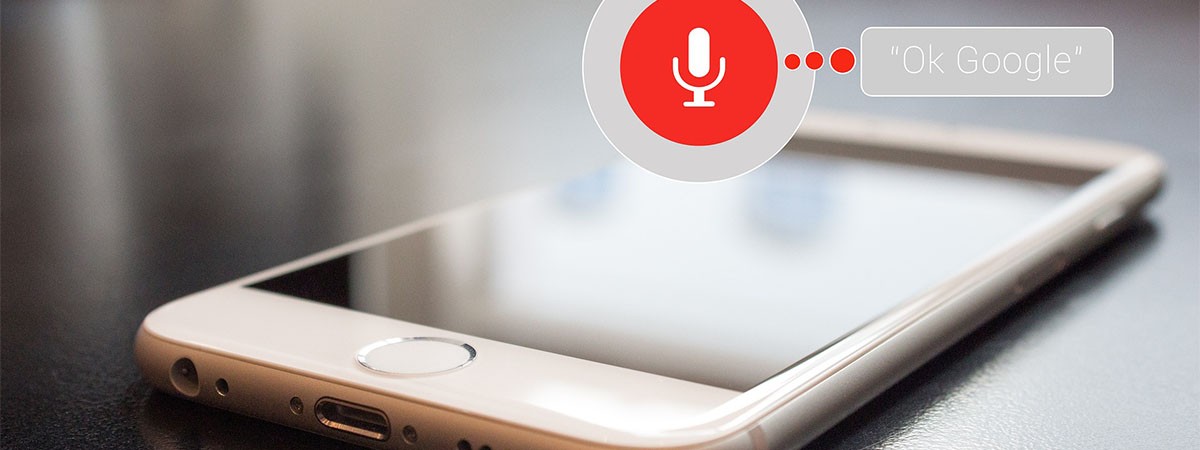 Ein weißes Smartphone liegt auf dem Tisch. Ein Mikrophonsymbol für die Aktivierung der Sprachsteuerung ist zu sehen.