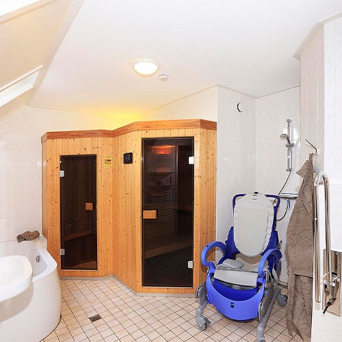 Das Badezimmer einer Ferienunterkunft in einem Park in Holland. Die Urlauber haben eine eigene Sauna im Bad sowie eine Badewanne und eine behindertengerechte Toilette. Unter der barrierefreien Dusche steht ein Rollstuhl, in dem der SMA-Patient gewaschen werden kann.