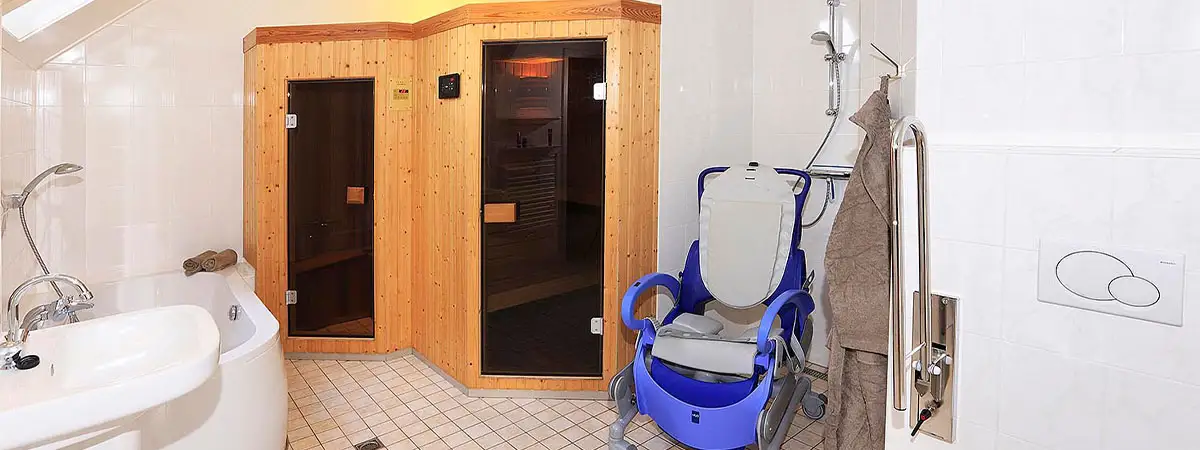 Das Badezimmer einer Ferienunterkunft in einem Park in Holland. Die Urlauber haben eine eigene Sauna im Bad sowie eine Badewanne und eine behindertengerechte Toilette. Unter der barrierefreien Dusche steht ein Rollstuhl, in dem der SMA-Patient gewaschen werden kann.
