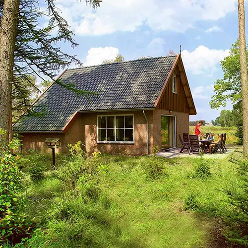 Ein Haus in einer Ferienanlage in den Niederlanden. Auf der Terasse sitzen Menschen, die Urlaub machen. Um das Haus herum ist es sehr grün aufgrund von Wiesen und Bäumen.