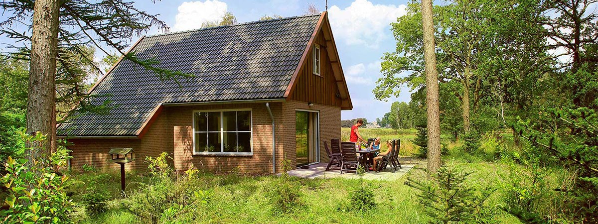 Ein Haus in einer Ferienanlage in den Niederlanden. Auf der Terasse sitzen Menschen, die Urlaub machen. Um das Haus herum ist es sehr grün aufgrund von Wiesen und Bäumen.