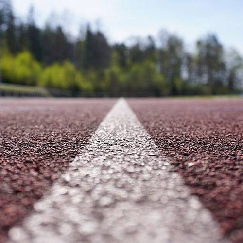 Eine weiße Linie auf einer roten Tartan-Bahn zum Joggen und Sport machen. Im Hintergrund sind grüne Bäume und ein blauer Himmel.