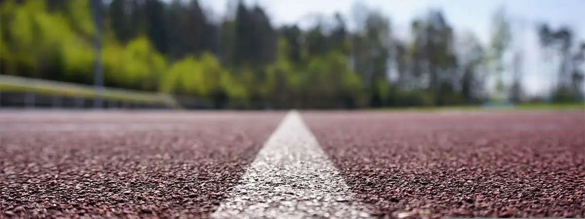 Eine weiße Linie auf einer roten Tartan-Bahn zum Joggen und Sport machen. Im Hintergrund sind grüne Bäume und ein blauer Himmel.