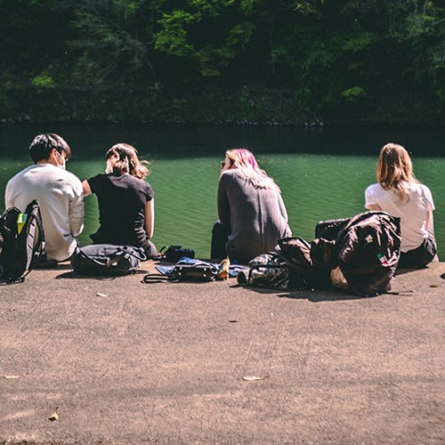 Eine Gruppe von Freunden verbringt ihre Freizeit gemeinsam an einem See. Die Jugendlichen sitzen in der Sonne und schauen aufs Wasser.