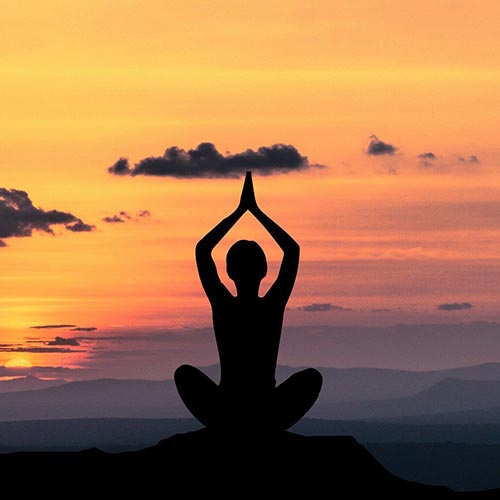 Eine Person meditiert oder macht Yoga. Es ist ihre Silhouette zu sehen. Sie sitzt im Schneidersitz und hat die Hände über dem Kopf. Im Hintergrund geht die Sonne über den Bergen auf/unter.
