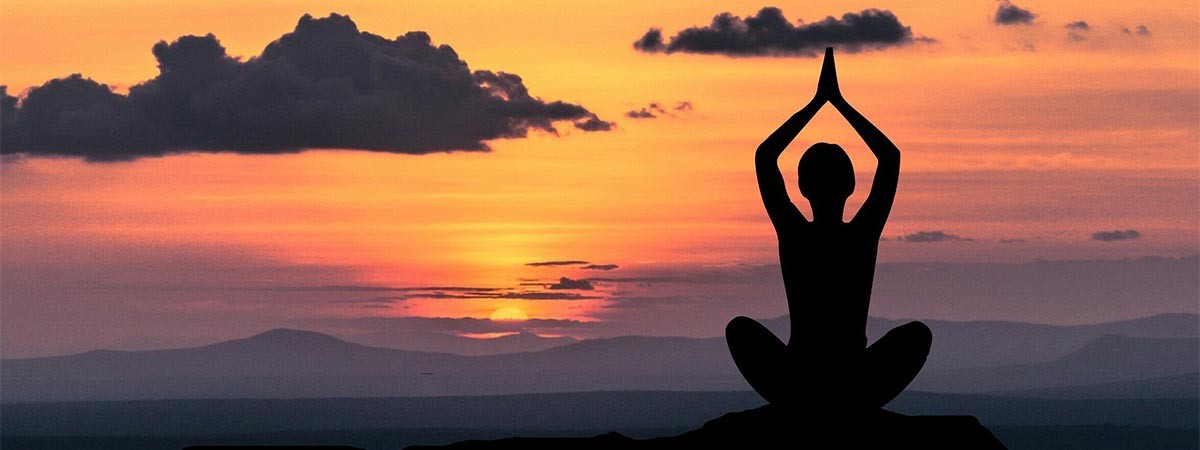 Eine Person meditiert oder macht Yoga. Es ist ihre Silhouette zu sehen. Sie sitzt im Schneidersitz und hat die Hände über dem Kopf. Im Hintergrund geht die Sonne über den Bergen auf/unter.