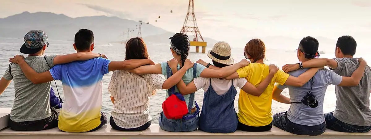 Acht Freunde sitzen Arm in Arm nebeneinander und blicken auf das Wasser. Die jungen Menschen halten sich gegenseitig unterstützend fest.